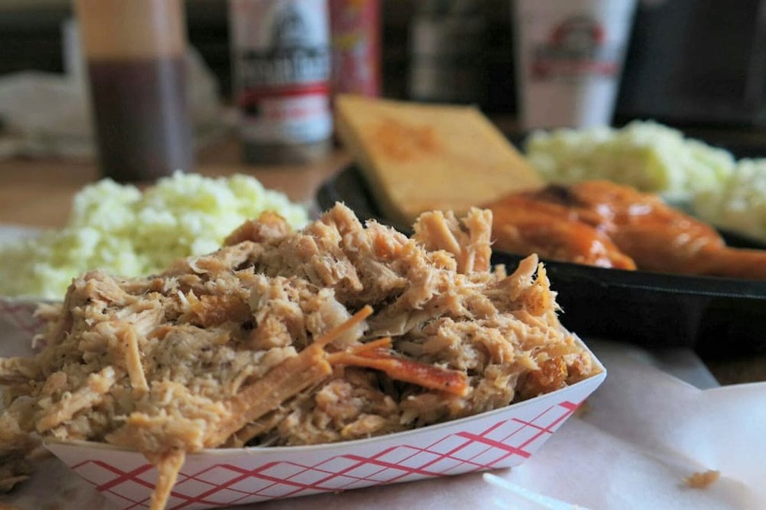 A serving of pulled pork is in a paper tray, with sides of coleslaw, cornbread, and sauced chicken wings on a black plate in the background. Bottles and cups are also visible in the blurred background.