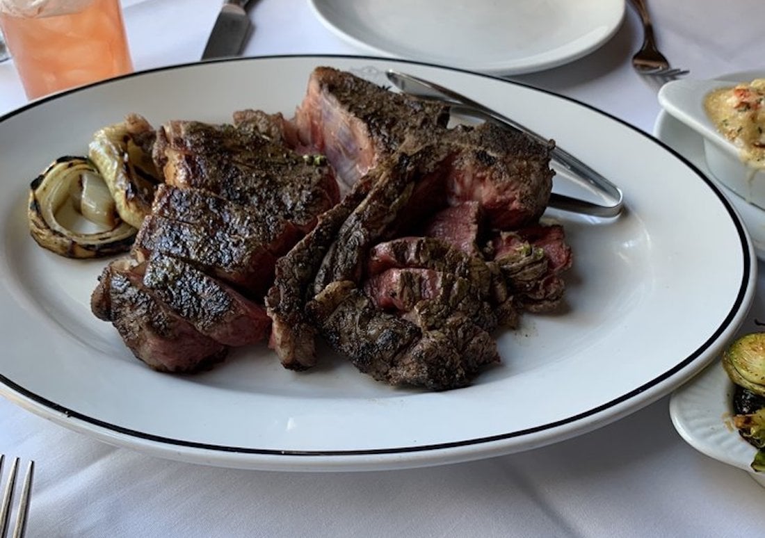A large, grilled steak sliced on a white oval plate with grilled onions on the side, set on a white tablecloth with a knife, fork, and side dishes visible nearby.