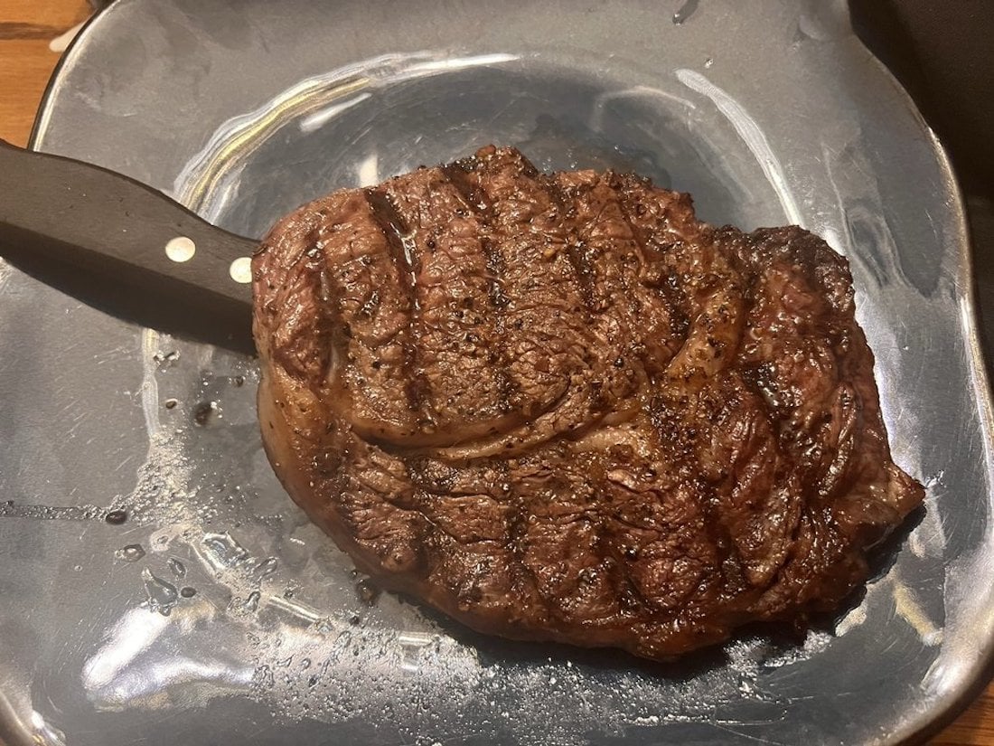 A cooked, seasoned steak with grill marks sits on a square metallic plate, with a steak knife partially under one side of the meat.