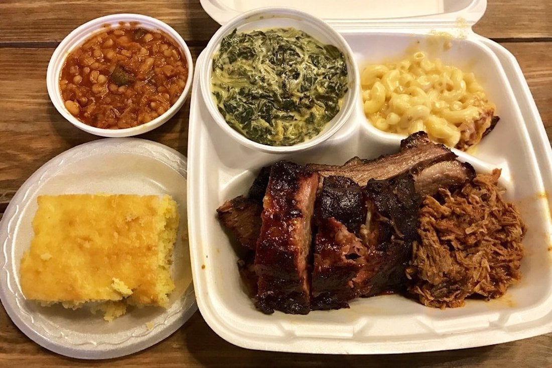 A takeout meal with baked beans, creamed spinach, macaroni and cheese, cornbread, and a foam container holding ribs, pulled pork, and brisket on a wooden table.