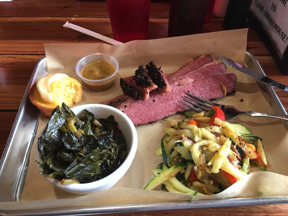 A tray with sliced brisket, cornbread, a cup of mustard sauce, sautéed greens in a bowl, and a side of mixed sautéed vegetables, served on parchment paper. Forks rest on the brisket.