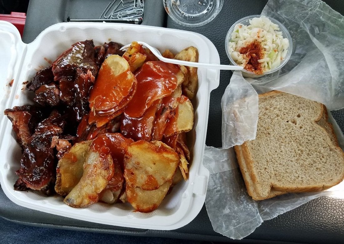 Barbecue meal in a foam container with sauced meat and sliced potatoes, alongside a small cup of coleslaw with garnish, and two slices of sandwich bread on wax paper. A plastic fork rests on the food.