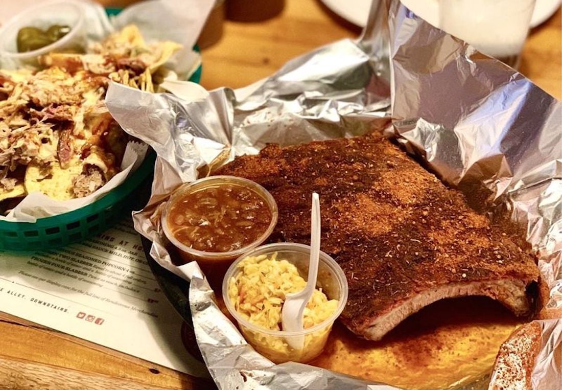 A tray with seasoned barbecue ribs on foil, served with small containers of baked beans and rice, next to a basket of nachos topped with shredded meat and cheese.