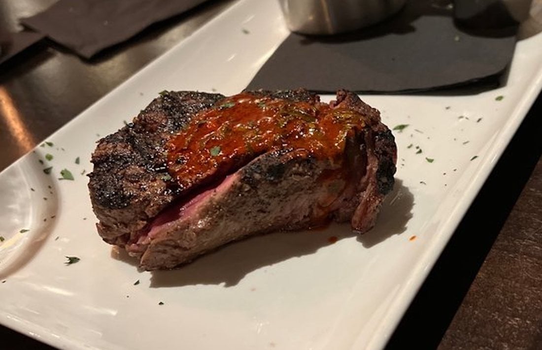 A grilled steak topped with red sauce sits on a white rectangular plate, garnished with chopped herbs. The steak appears juicy with a medium-rare center.
