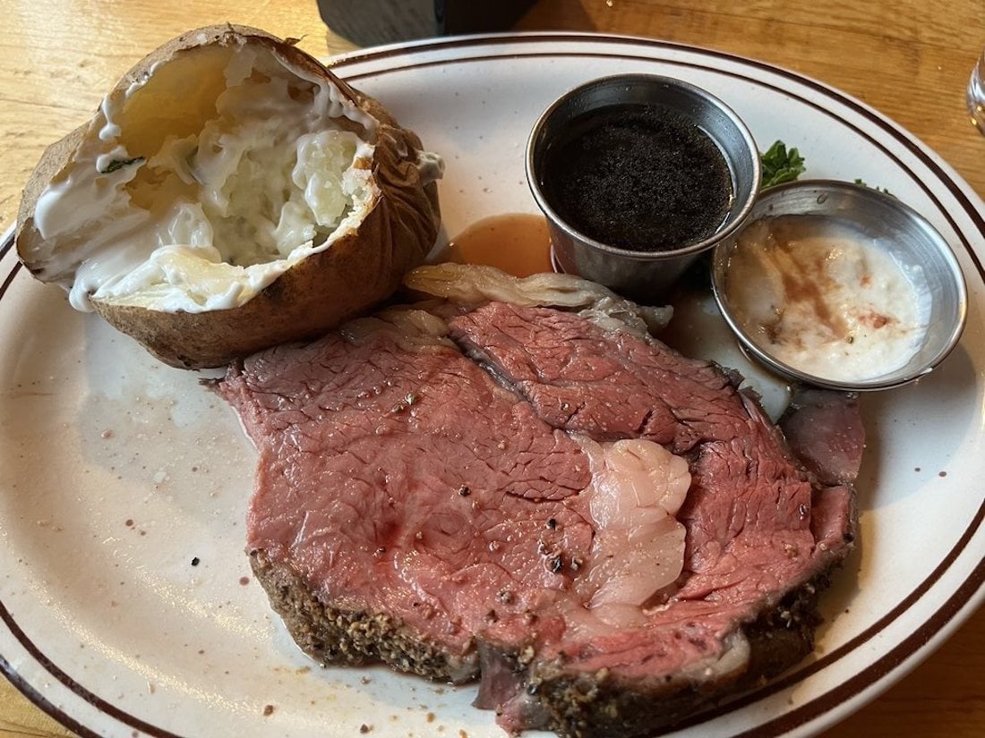A plate of prime rib with visible marbling, served with a baked potato topped with sour cream, and small cups of au jus, horseradish sauce, and a dark seasoning or condiment on a wooden table.