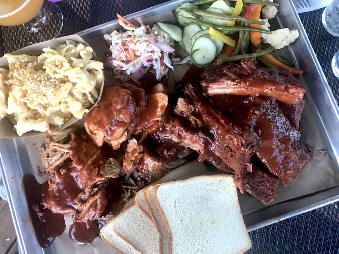 A tray of barbecue food with sauced ribs, pulled pork, macaroni and cheese, coleslaw, pickled vegetables, and slices of white bread, arranged on brown paper.