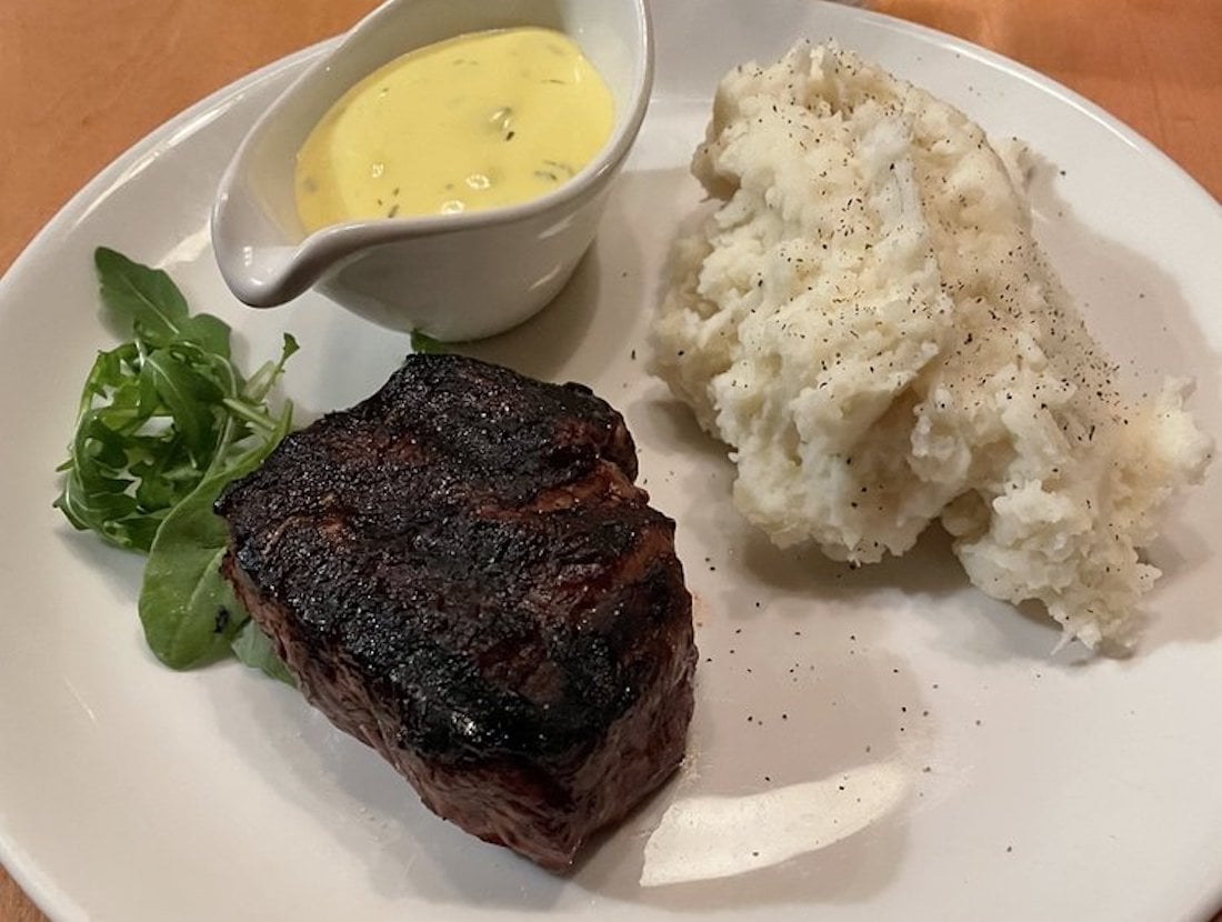 A white plate with a grilled steak, a portion of mashed potatoes topped with black pepper, a small cup of creamy yellow sauce, and a sprig of fresh greens on the side.