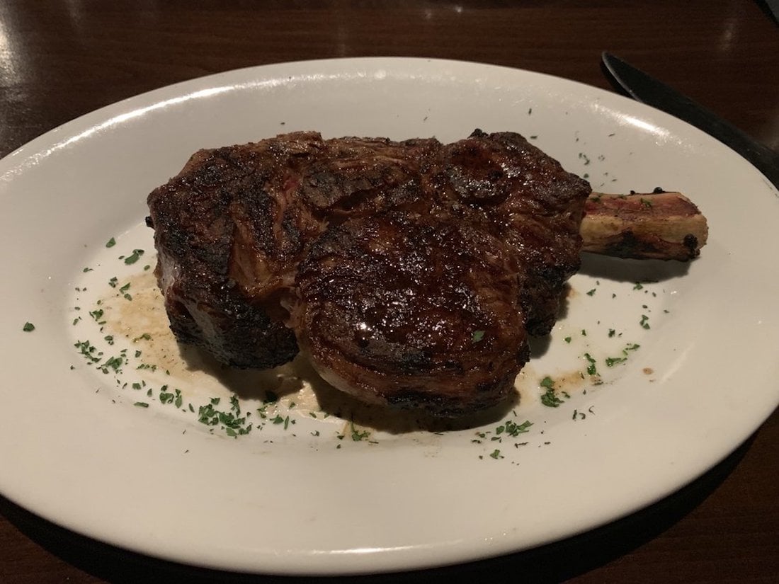A grilled bone-in ribeye steak served on a white plate, garnished with chopped herbs, sits on a dark wooden table.