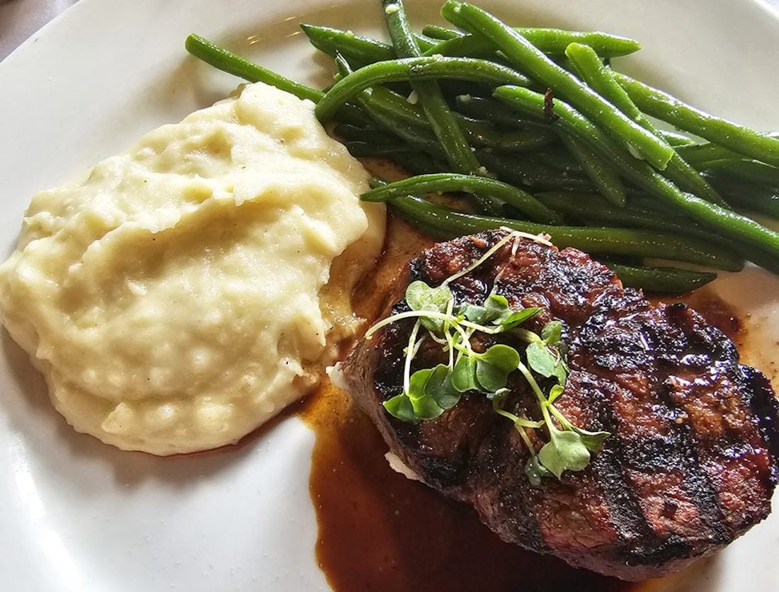 A plate with grilled steak topped with microgreens, a serving of creamy mashed potatoes, and sautéed green beans on the side.