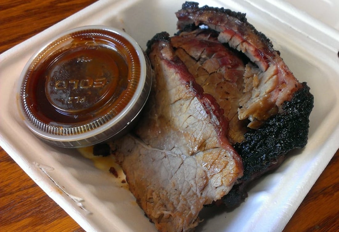 Two thick slices of smoked brisket with a dark bark edge are served in a white foam takeout container next to a small plastic cup of barbecue sauce.