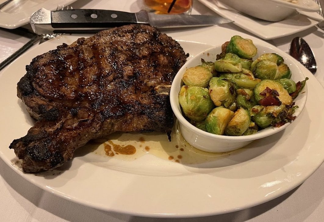 A grilled steak sits on a white plate next to a small bowl of roasted Brussels sprouts. A steak knife rests on the plate, and the meal is served on a white tablecloth.