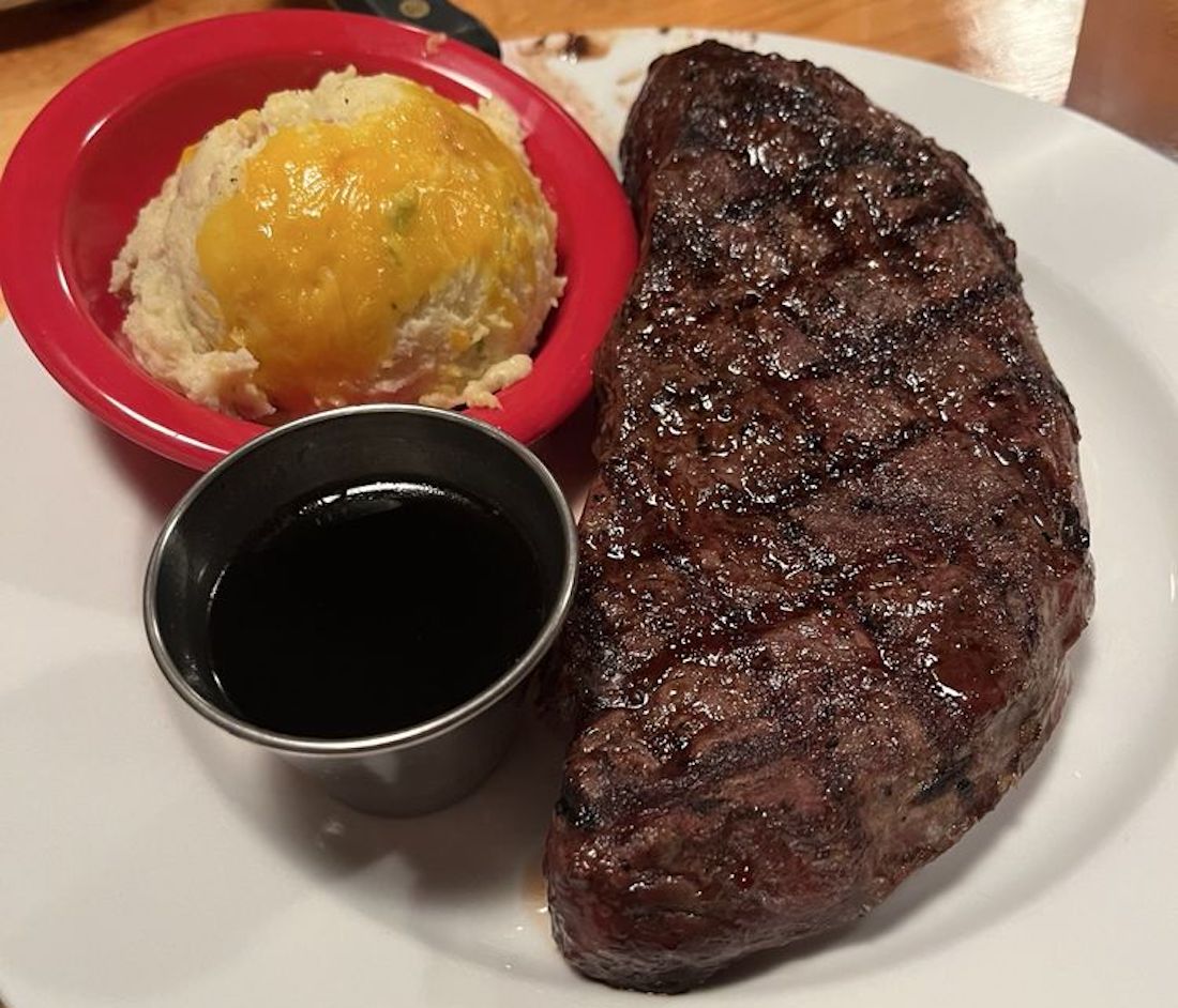A grilled steak with sear marks is served on a white plate alongside a scoop of mashed potatoes topped with melted cheese and a small cup of dark dipping sauce.