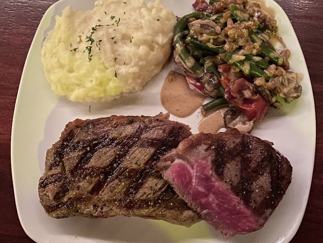 A plate with a grilled steak cooked medium rare, mashed potatoes topped with herbs, and a side of sautéed green beans, mushrooms, and tomatoes in a creamy sauce.