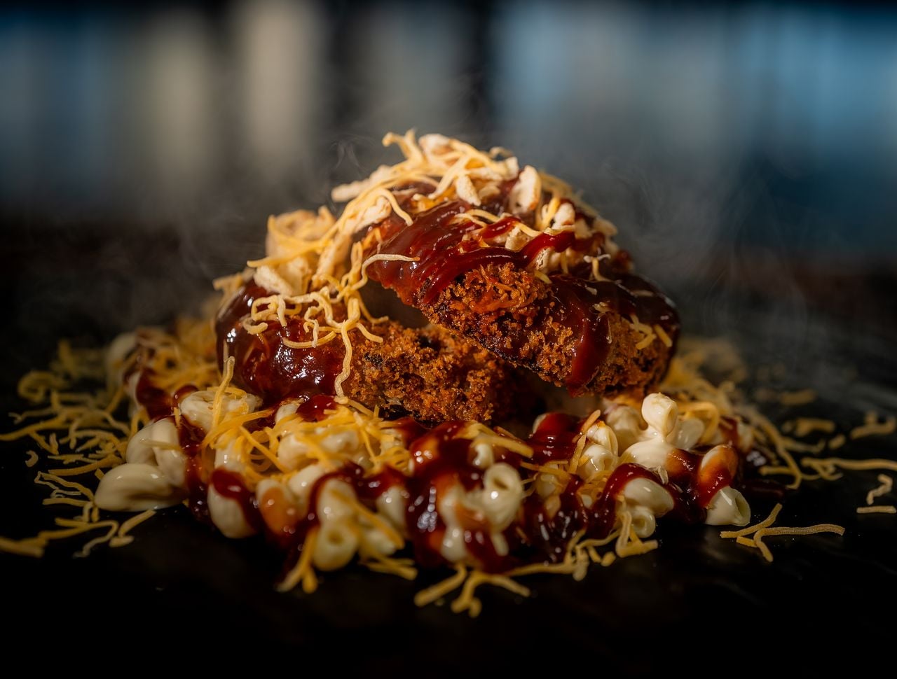 Close-up of a serving of macaroni and cheese topped with two slices of meatloaf, covered in barbecue sauce and shredded cheese, with steam rising from the hot dish.