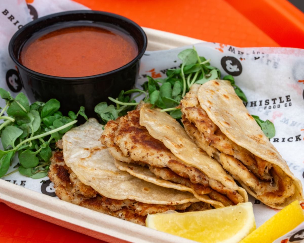 Three folded birria tacos with grilled tortillas are arranged on a tray, served with fresh greens, a lemon wedge, and a cup of reddish dipping sauce on the side.