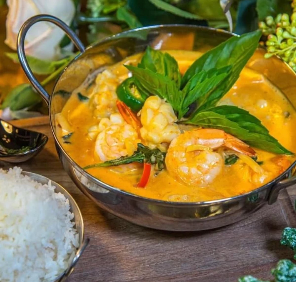 A metal bowl of creamy shrimp curry soup, garnished with fresh basil leaves and surrounded by green herbs, sits on a wooden table next to a serving of steamed white rice—worthy of being the best soup in every state.