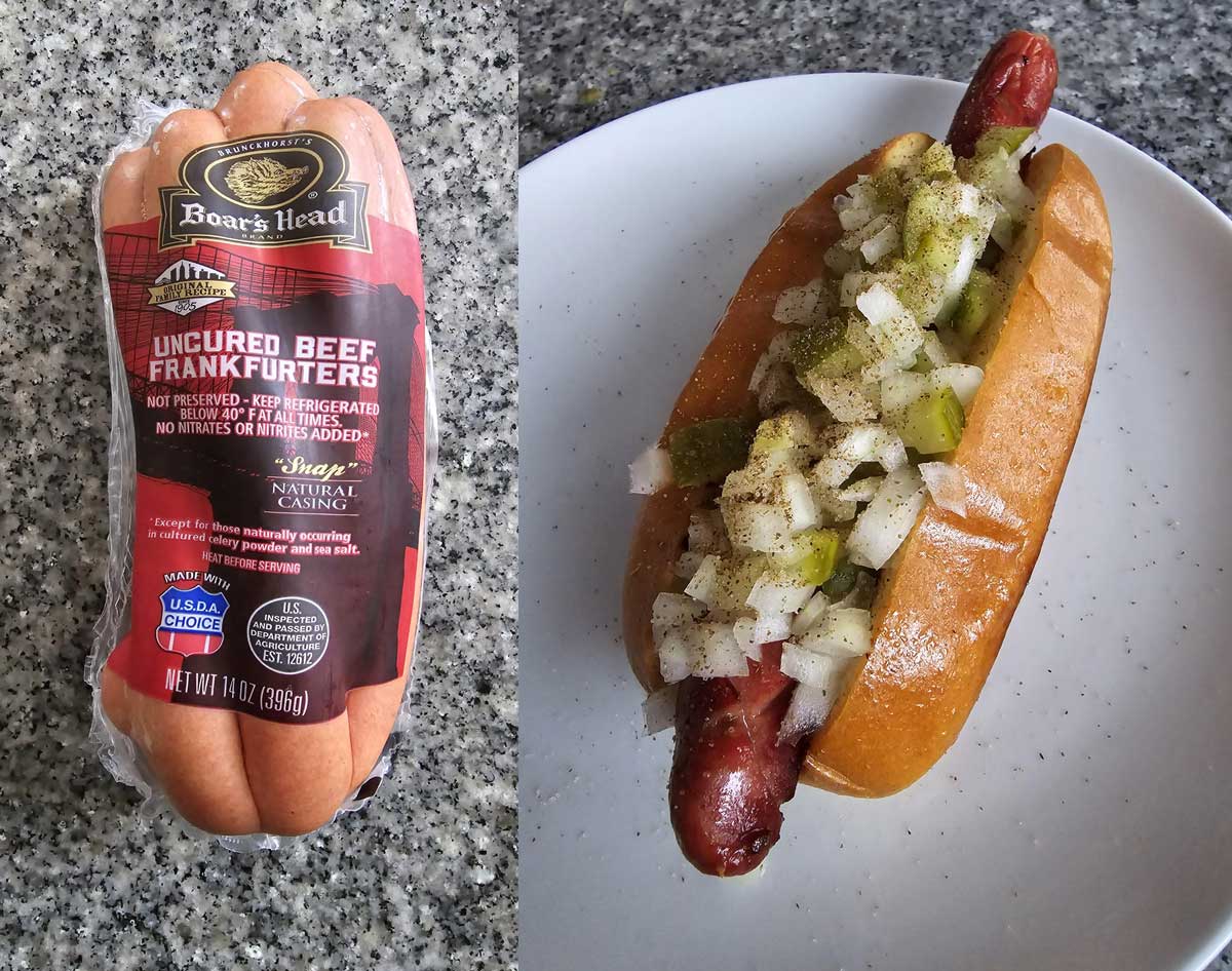 A pack of Boar's Head uncured beef frankfurters sits on a gray countertop next to a plate with a cooked hot dog in a bun topped with chopped onions, pickles, and black pepper.