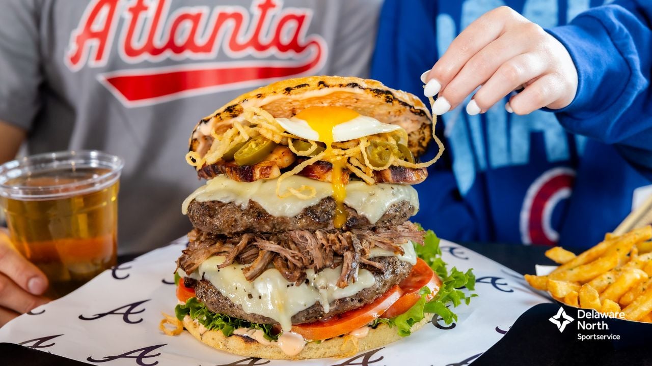 A close-up of a large stacked burger with cheese, pulled meat, lettuce, tomato, fried onions, jalapeños, and a fried egg on top, next to fries and a drink. Two people in sports shirts sit behind it.