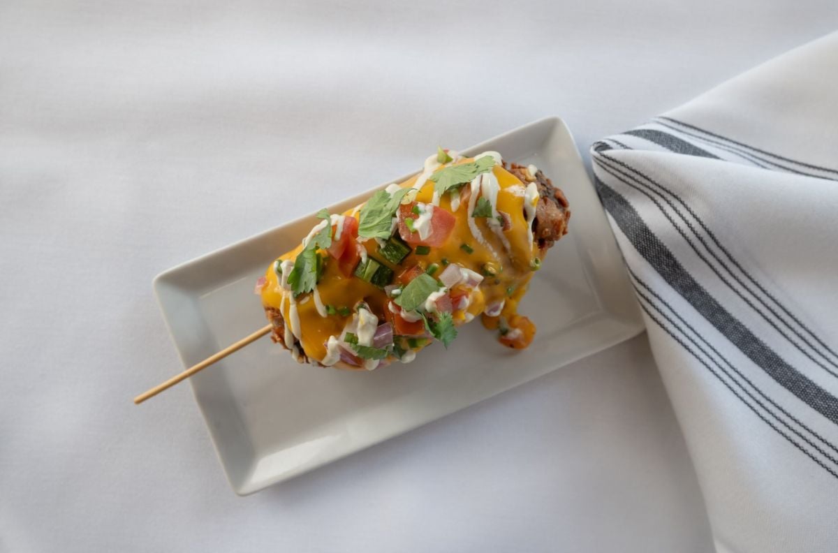 A corn dog on a rectangular white plate, topped with melted cheese, diced tomatoes, onions, and green herbs, next to a white napkin with black stripes on a white tablecloth.