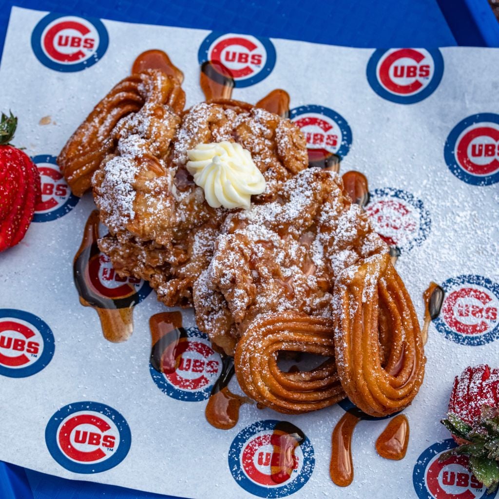 A Mickey Mouse-shaped churro dessert with powdered sugar, a swirl of whipped cream, and syrup, served on Cubs-branded paper. Two strawberries are placed on the side, all on a blue surface.