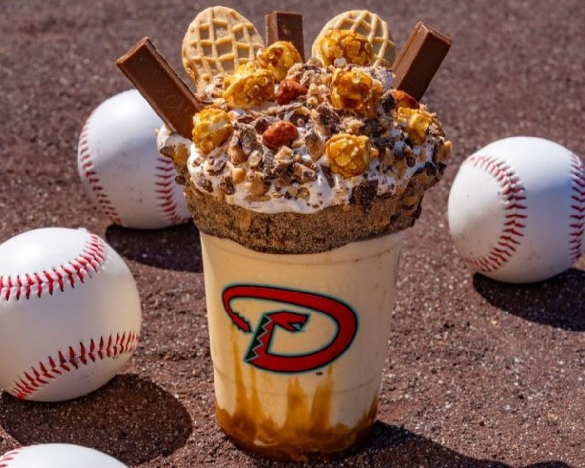 A milkshake in a cup with a "D" logo, topped with mini waffles, chocolate bars, popcorn, nuts, and crushed cookies, surrounded by four baseballs on a dirt surface.