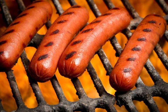 Four grilled hot dogs with char marks are cooking on a metal grill over an open flame, with orange fire glowing beneath the grill grates.
