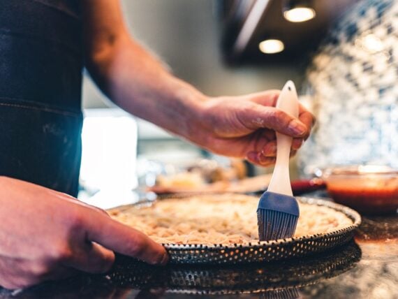 A person uses a blue silicone brush to prepare a pizza crust on a round baking tray, with ingredients and sauce visible in the background on a kitchen counter.