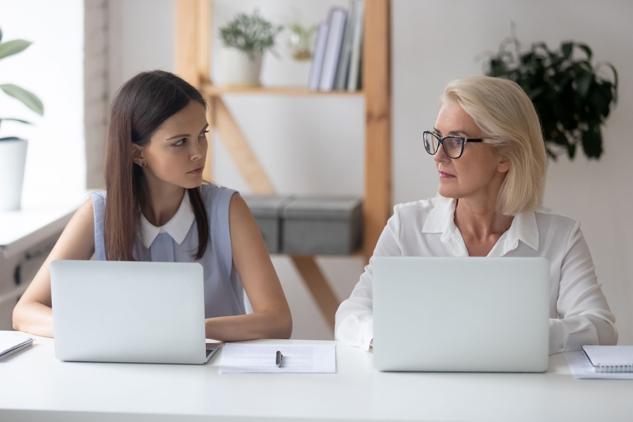 Different ages women colleagues 50s and young female workmates sit at office desk looking with enmity at each other. Negative attitude, conflict at workplace, misunderstanding generational gap concept