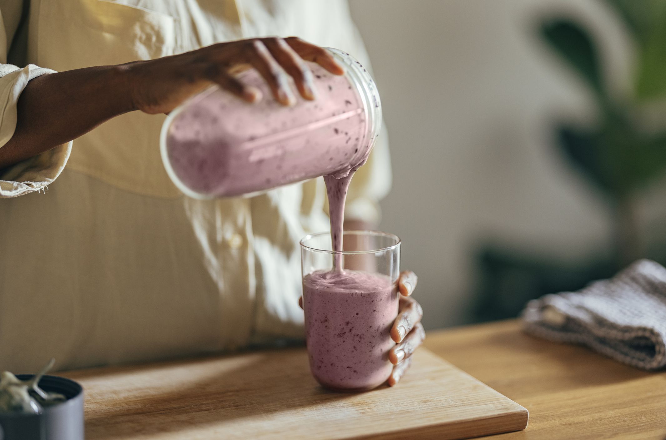 Close-up of hands of an Afro-American woman wearing beige shirt and pouring smoothie into a glass on a kitchen table. There is a part of a blender, a cutting board and a cloth on the desk in front of her. In the corner of a room sits a big plant.