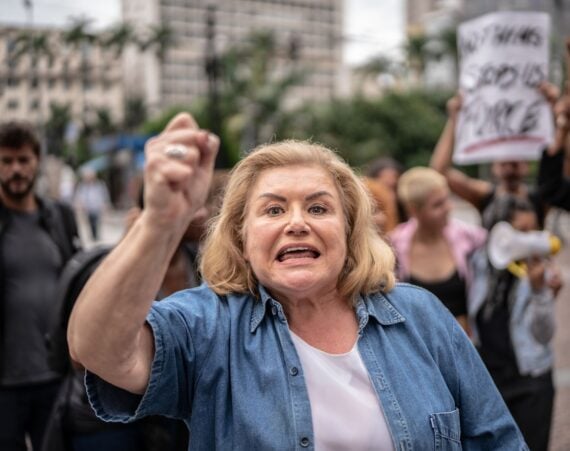 A woman with blonde hair raises her fist and shouts during a protest, with other people and signs visible in the blurry background.