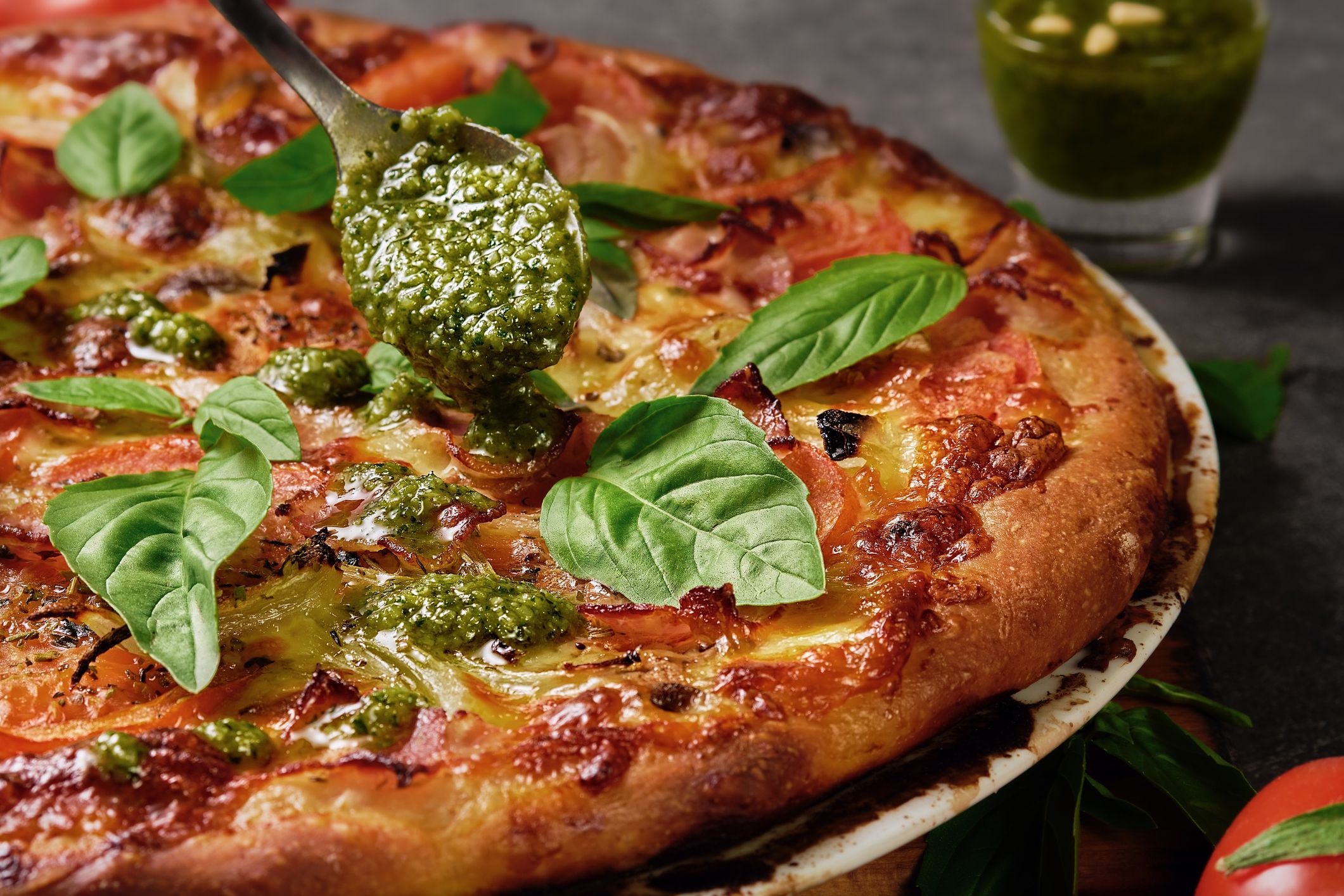 Homemade Italian pizza with ham, mozzarella, tomatoes and basil leaves on a dark background. Green pesto is added to the pizza. Close-up, selective focus.