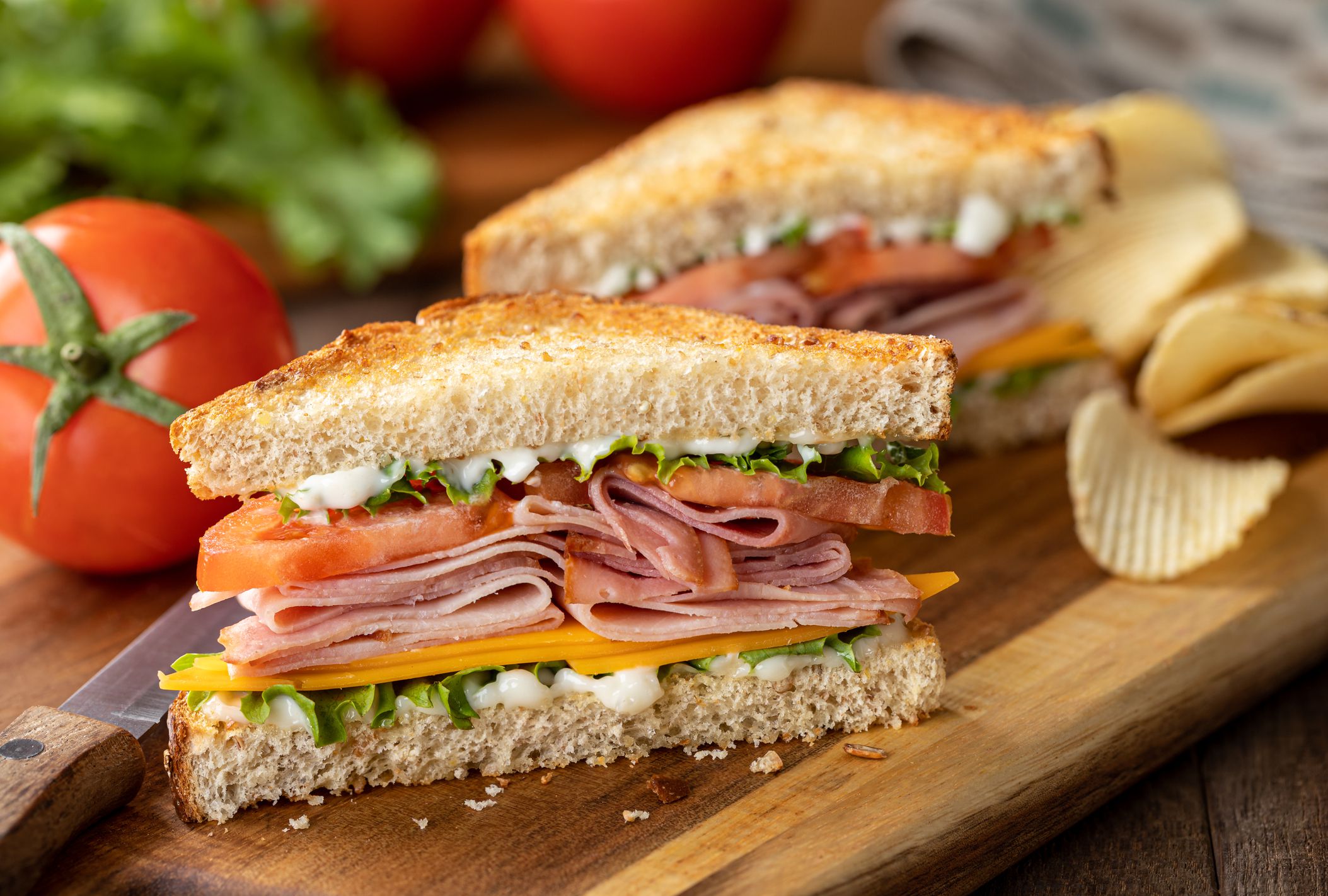 Ham sandwich with cheese, lettuce and tomato on whole grain bread. Lettuce and tomatoes in background
