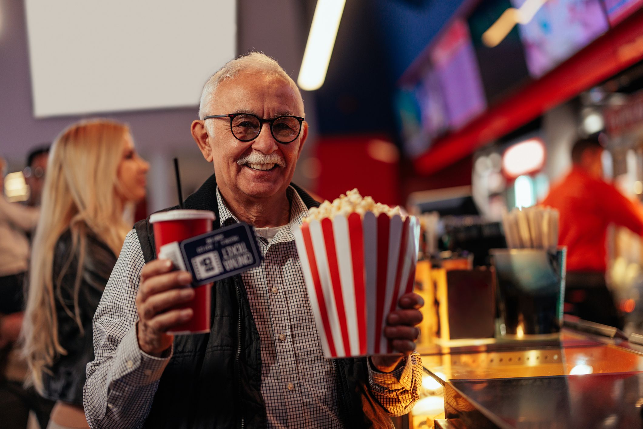 A cheerful senior man is at the movie theater, holding tickets, popcorn and soda in his hands and on his way to the projection hall.