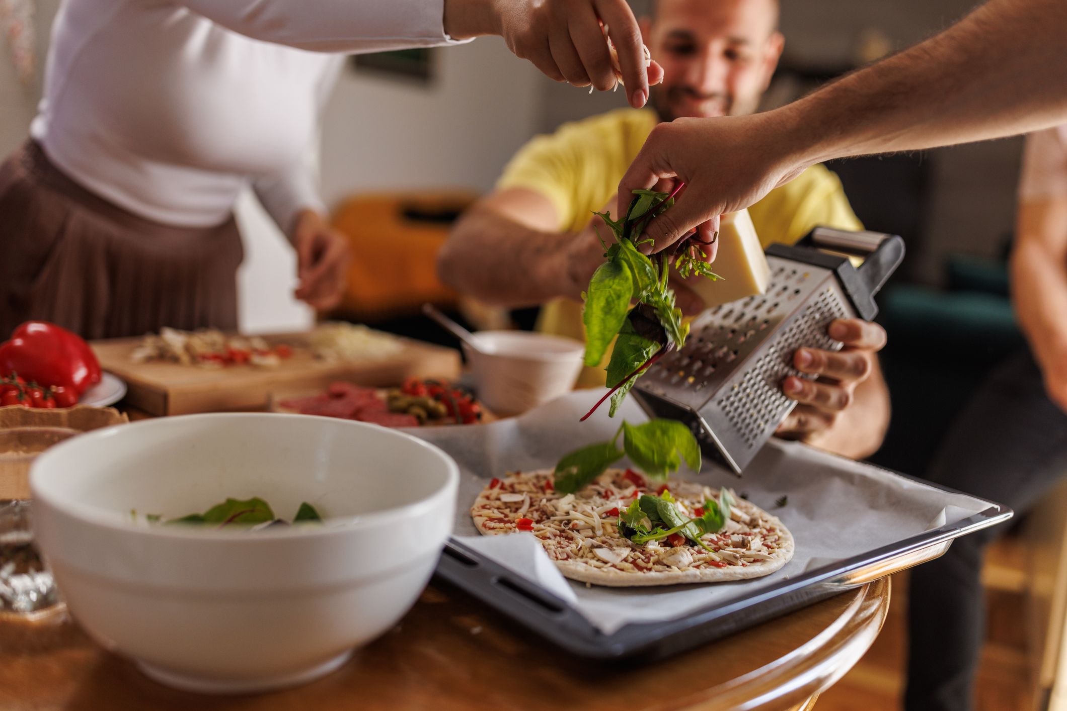 Close up shot of two unrecognizable friends sprinkling freshly grated cheese and greens on top of a homemade pizza.