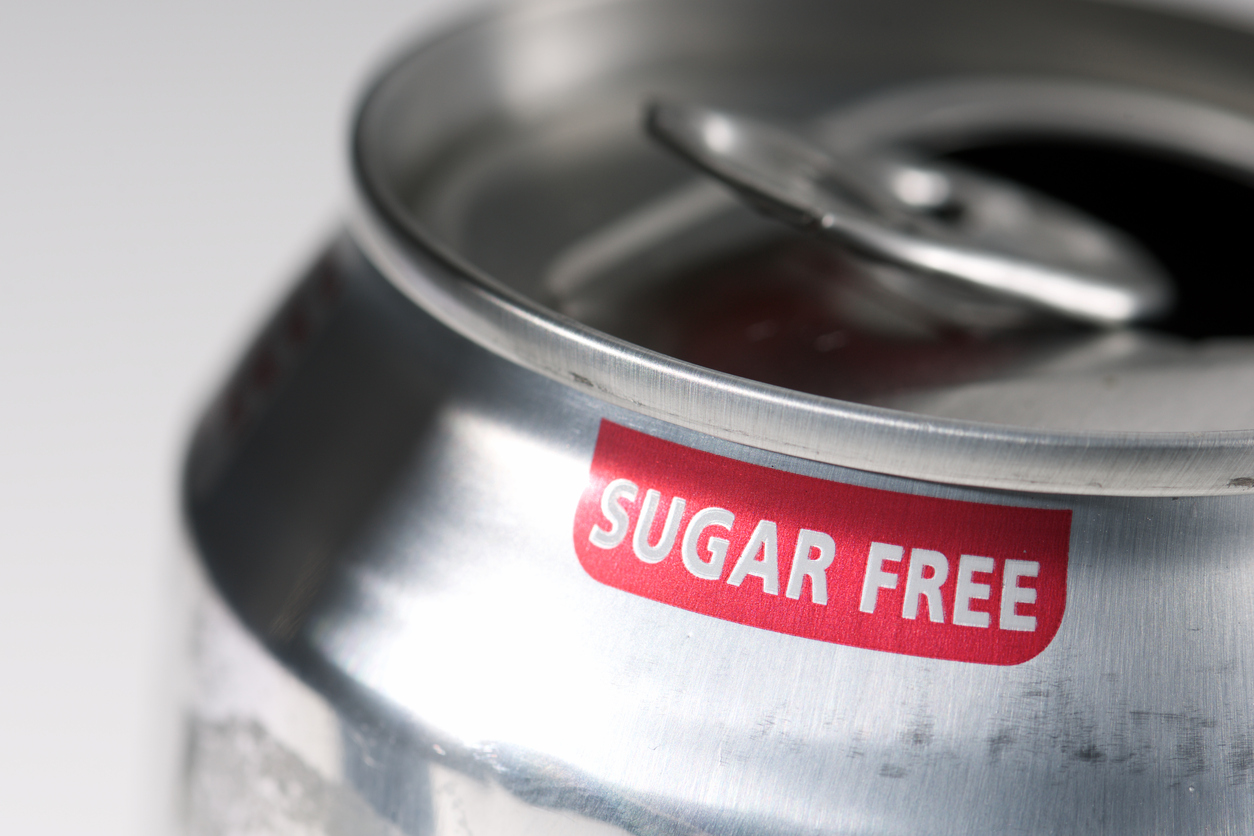 A close-up of an open silver soda can with a red label that reads "SUGAR FREE" near the rim. The background is out of focus and light in color.