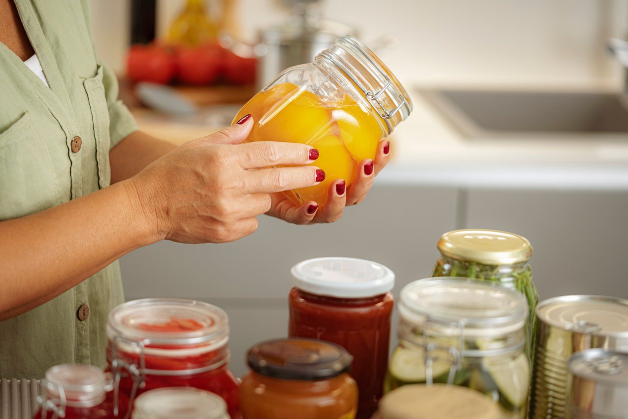 A person with red nail polish holds a jar of preserved yellow fruit in a kitchen. Various jars of homemade preserves, pickles, and sauces are arranged on the counter.