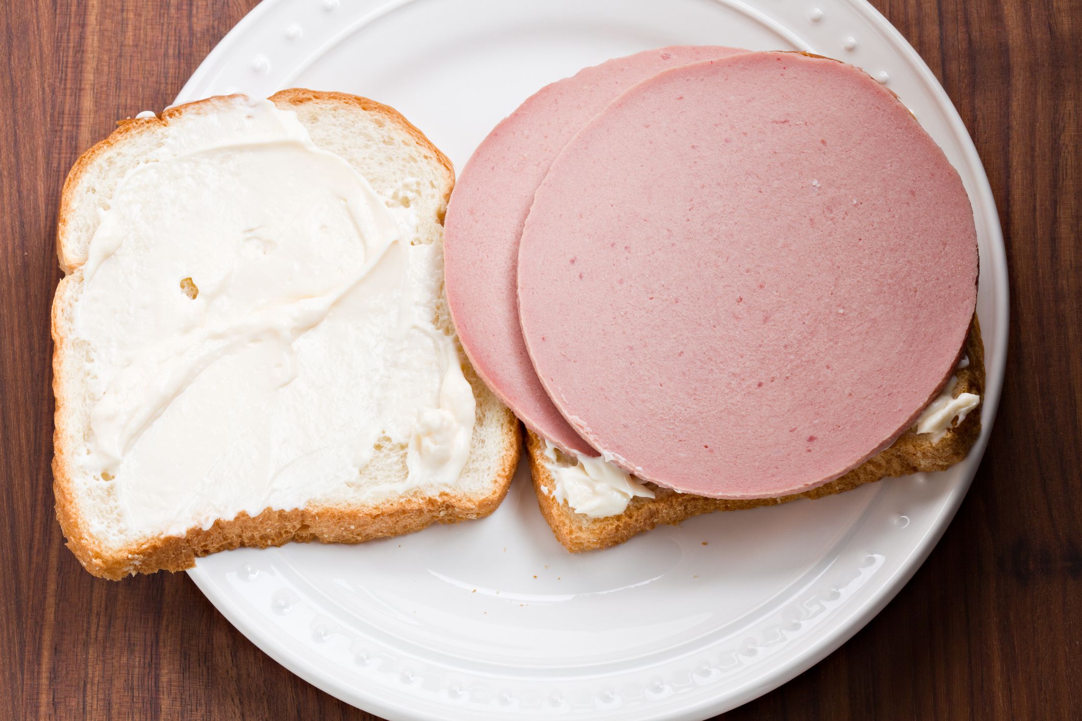 An overhead close up view of a white plate with an open face bologna sandwich on white sourdough bread.