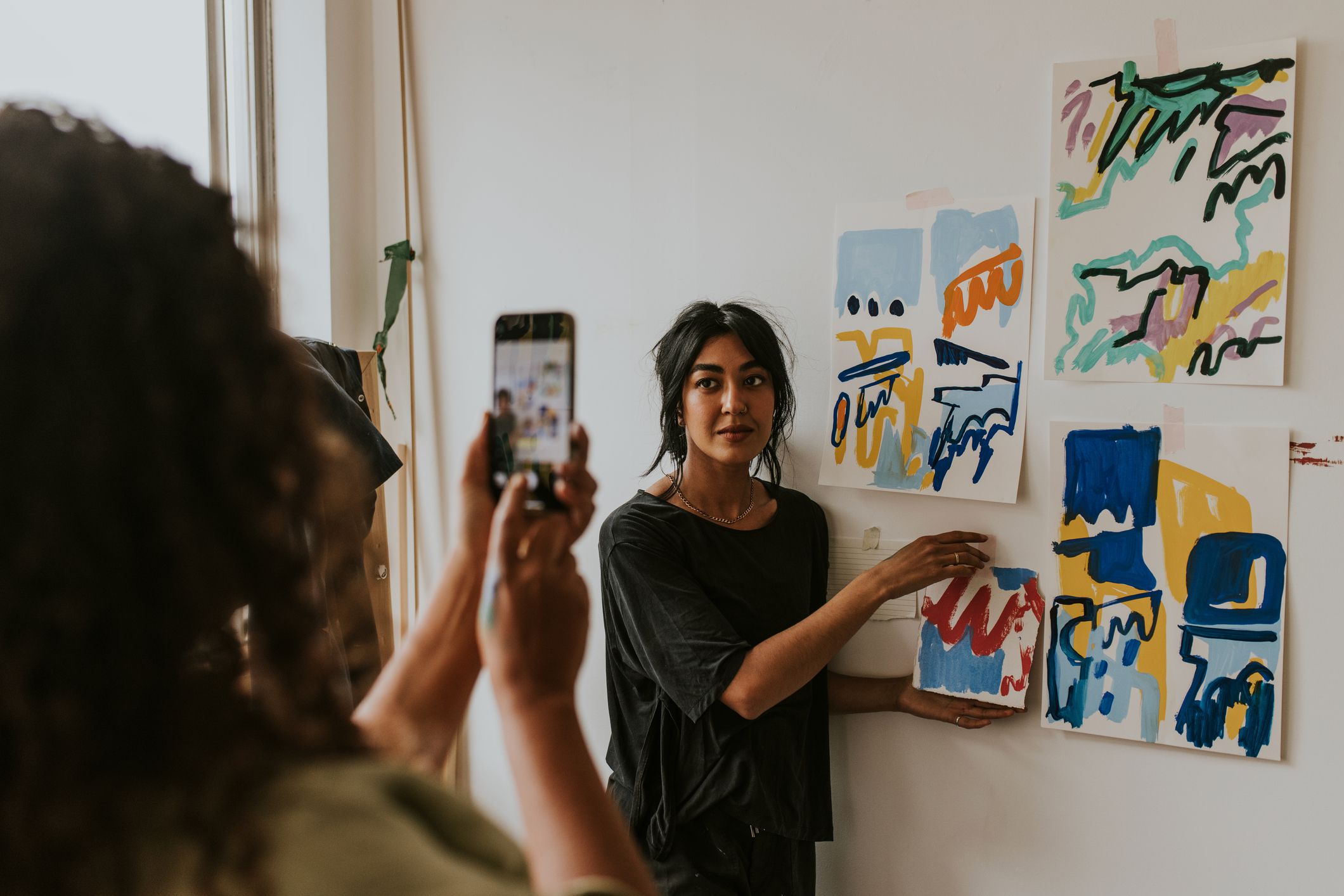 Friend taking a photo with phone camera of her artist friend by a mood board on a white wall