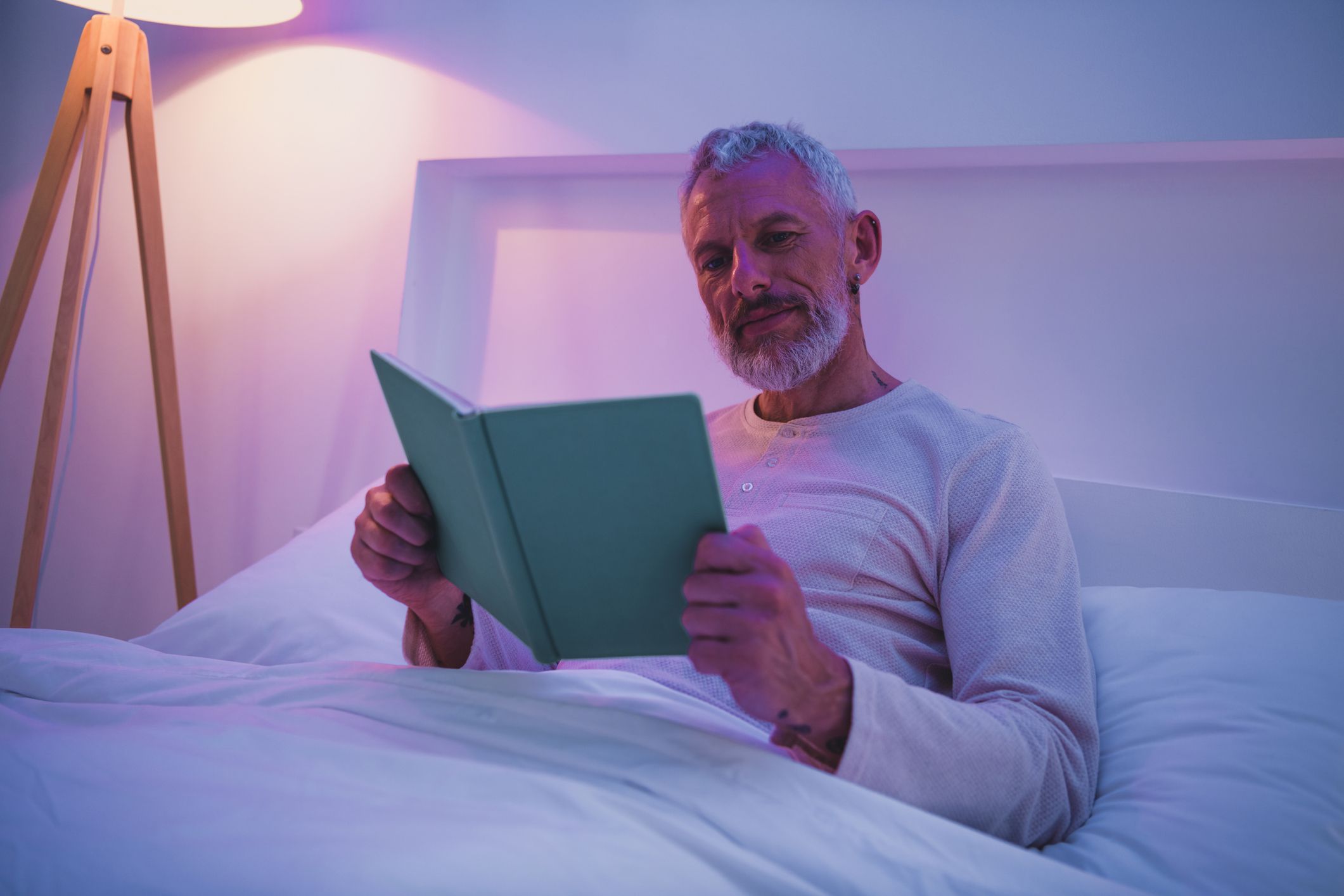 An elderly man with grey hair is reading in bed at home, surrounded by calming soft light, enjoying a peaceful evening in comfortable pajamas.