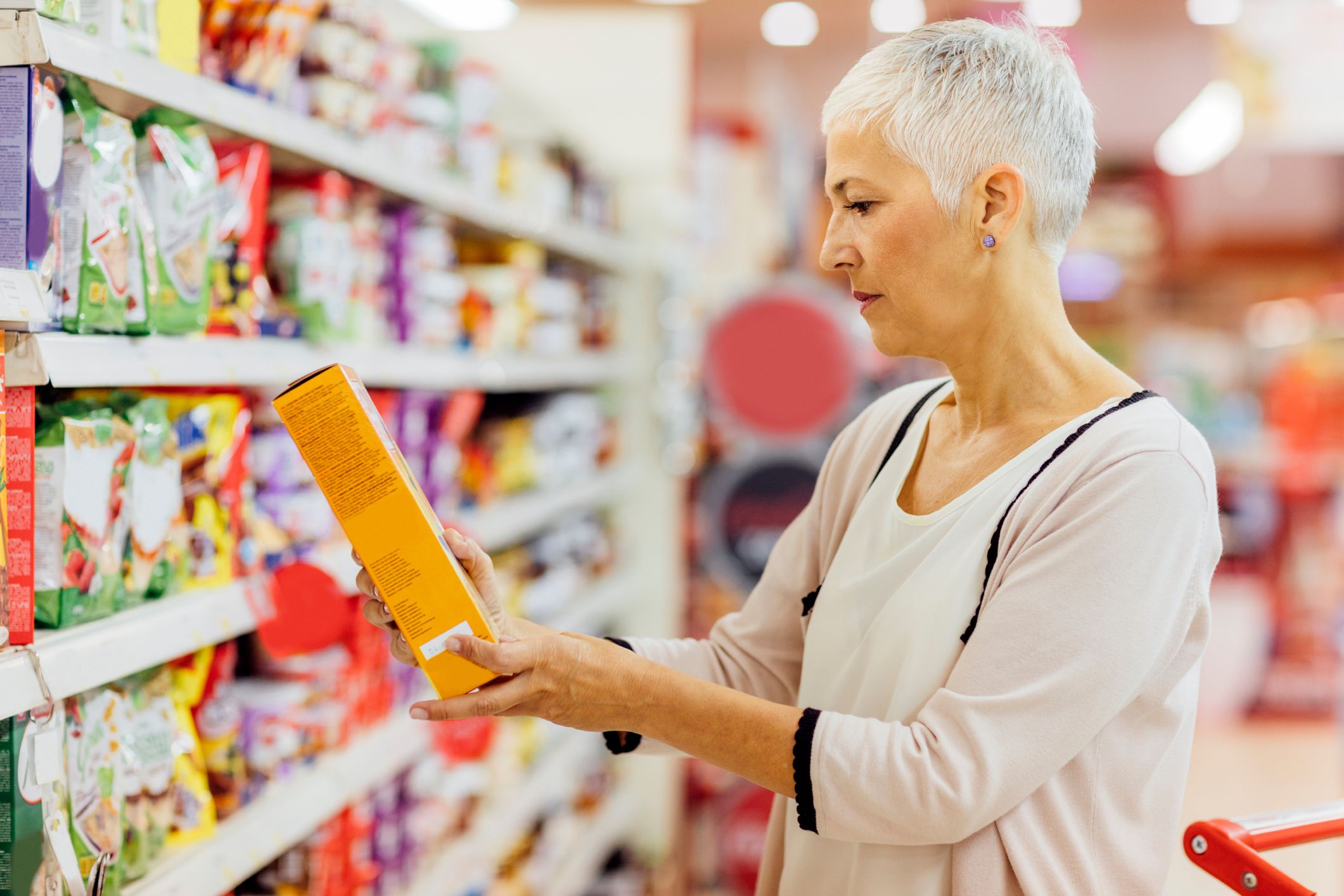 Mature woman shopping in local supermarket. Standing by produce stand and choosing cereals. Holding box and reading nutrition label.