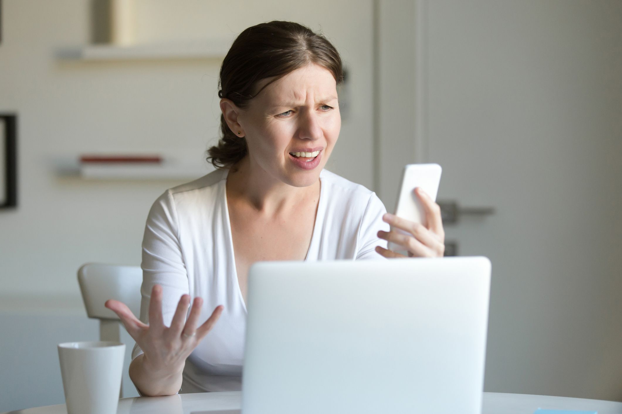 Portrait of a young woman at the desk with a laptop, looking puzzled at the screen of the mobile she holds at her hand and message. Business concept photo, lifestyle