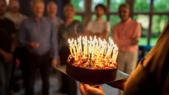 A person holds a chocolate birthday cake with many lit candles, while a group stands in the background, slightly out of focus—one of the fun warning signs you’re getting old.