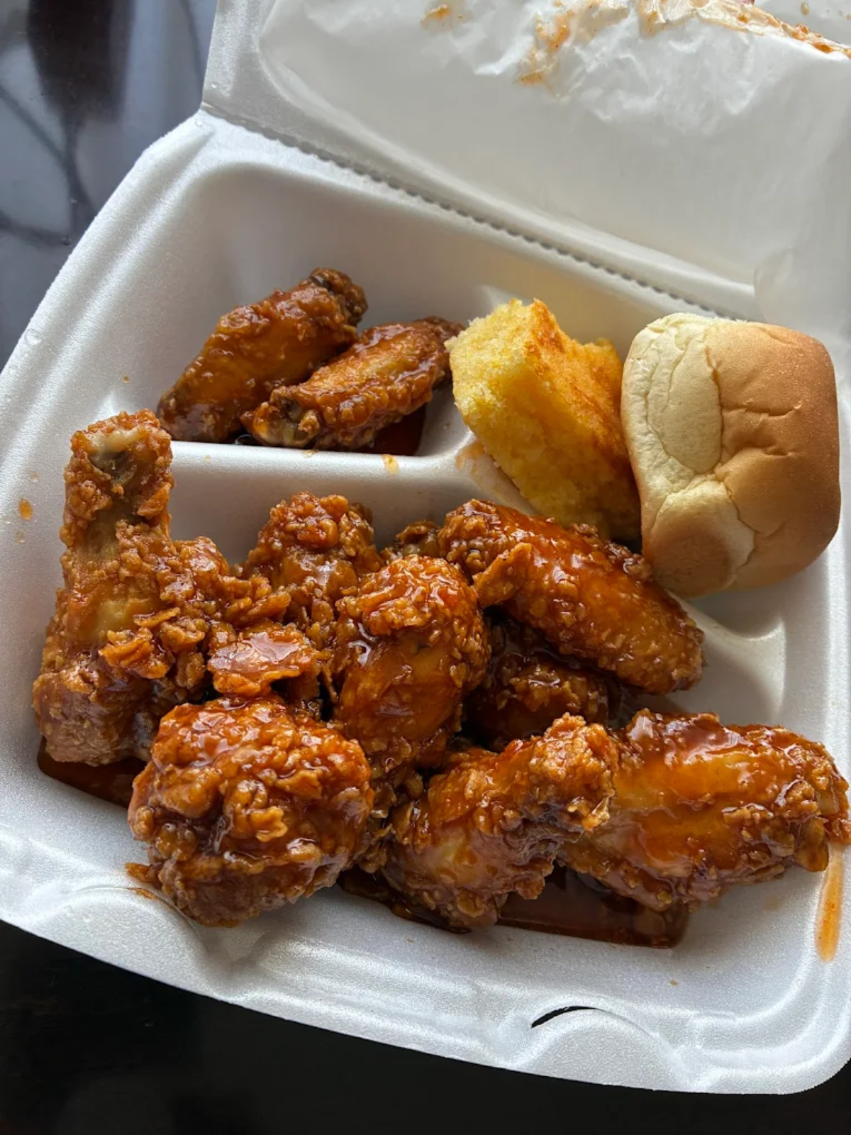 A styrofoam takeout container with saucy fried chicken wings, a piece of cornbread, and a dinner roll. The sectioned container keeps the food items separated.
