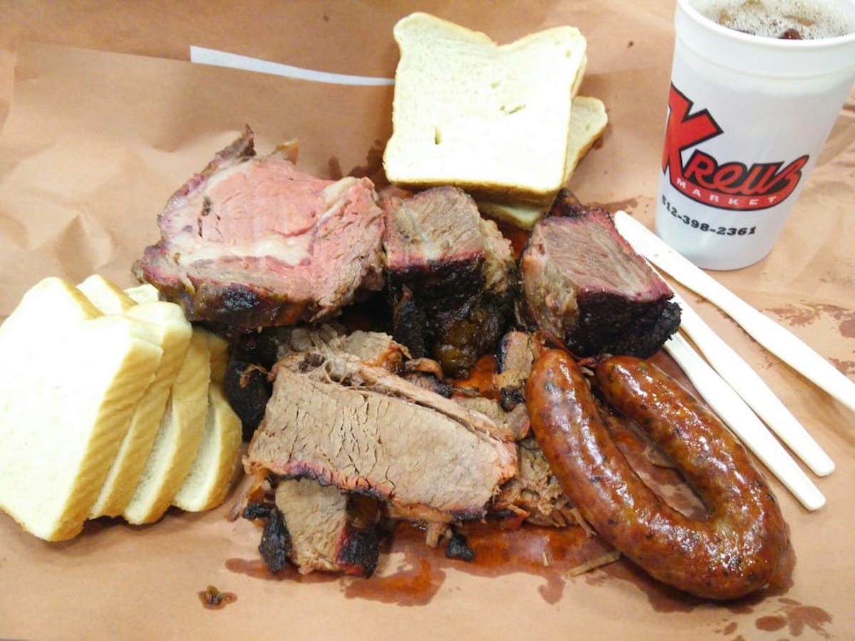 A platter of barbecue from one of the best hole-in-the-wall BBQ joints, featuring slices of white bread, brisket, sausage, and ribs on brown paper, beside a "Kreuz Market" drink cup and two white plastic utensils.