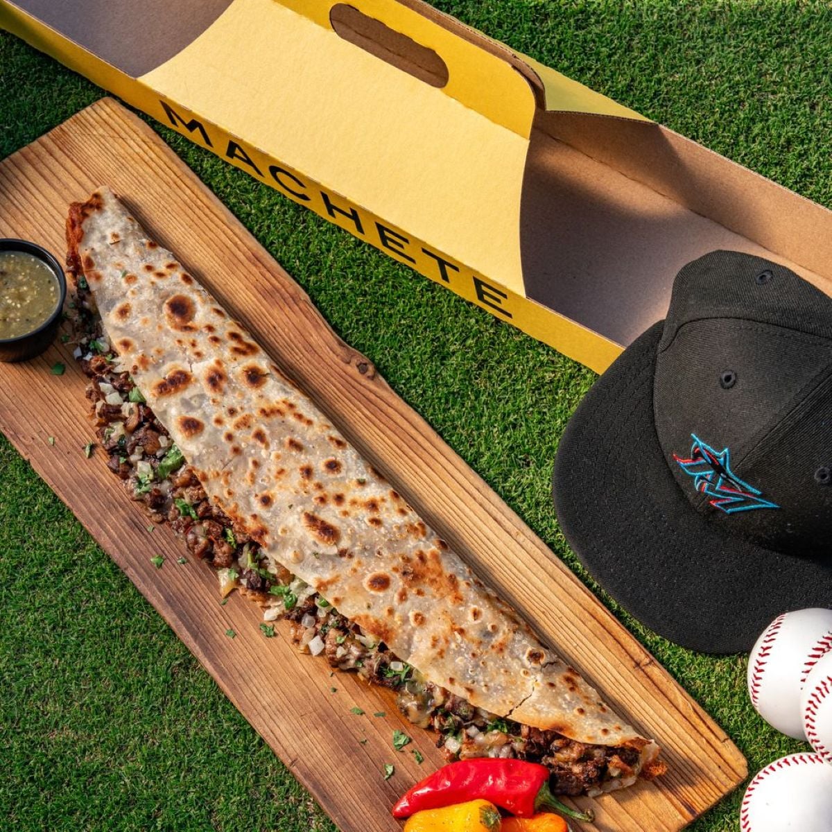 A long, stuffed quesadilla called "Machete" on a wooden board with salsa, chili peppers, a black Miami Marlins hat, two baseballs, and a yellow box on grass.