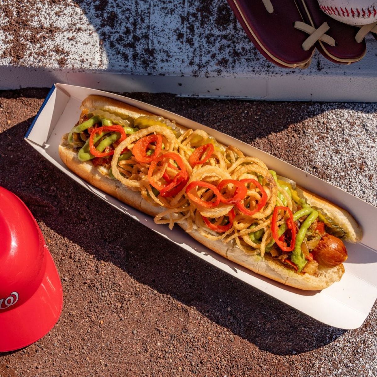 A long hot dog in a paper tray topped with crispy fried onions, sliced red peppers, and green relish, sitting on a dirt surface near a red baseball helmet and a pair of baseball gloves.