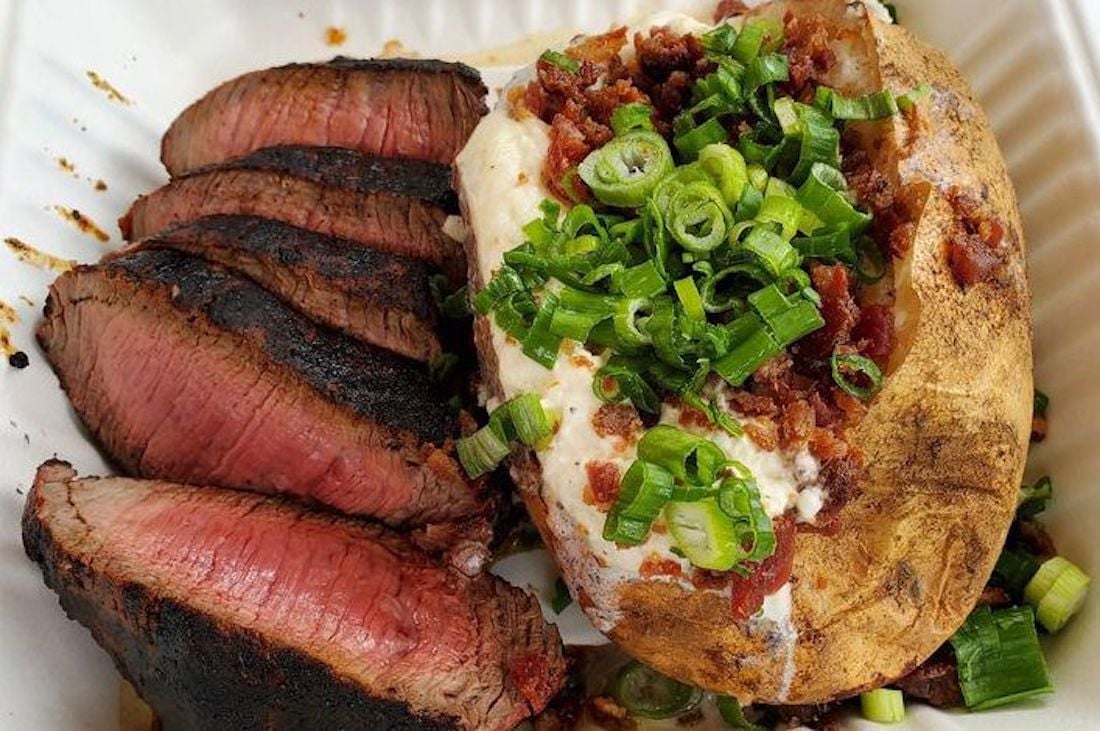 Sliced medium-rare steak next to a baked potato topped with sour cream, bacon bits, and chopped green onions on a white plate.