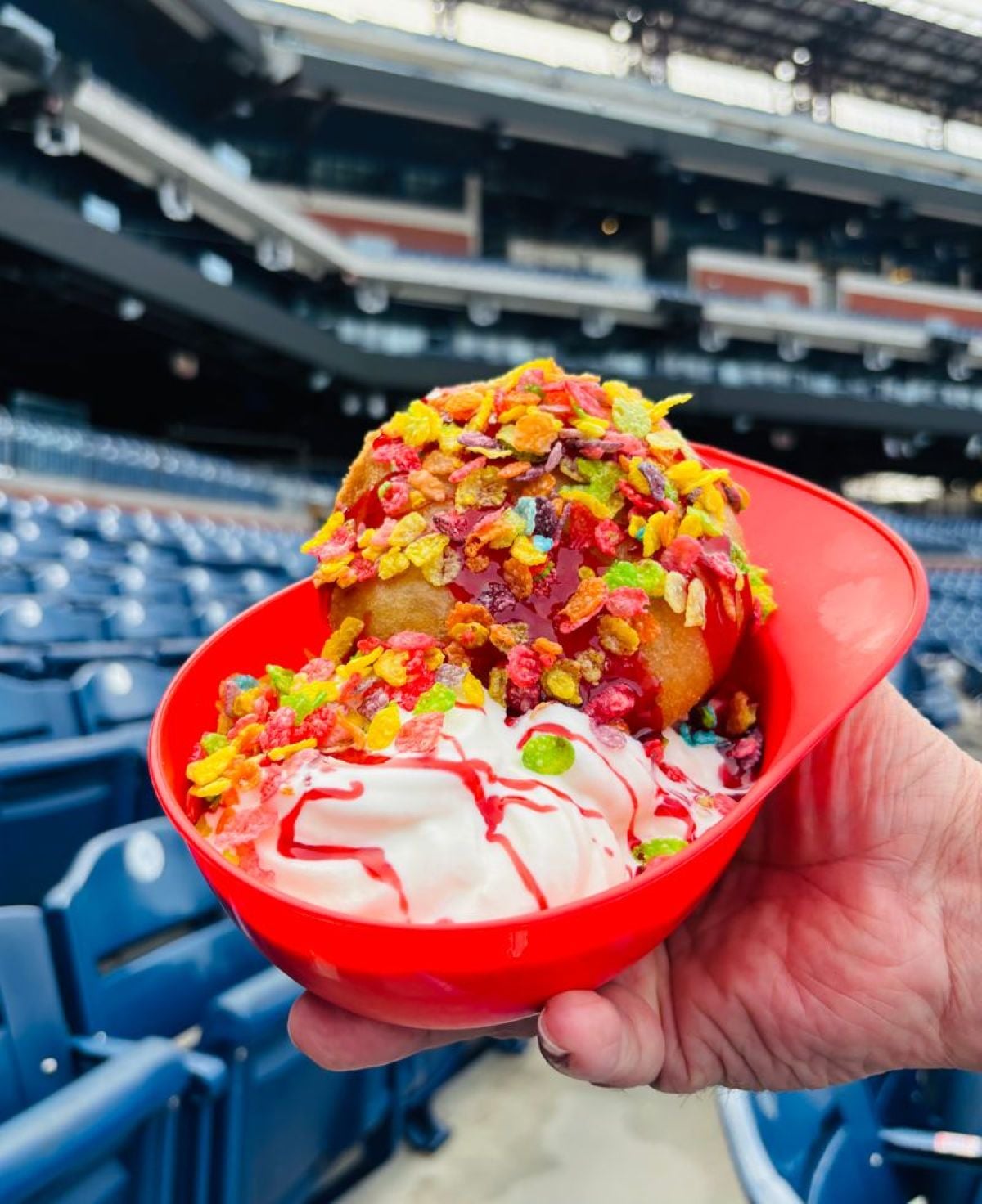 A hand holds a bright red plastic bowl shaped like a baseball helmet, filled with ice cream topped with whipped cream, berry syrup, and colorful Fruity Pebbles cereal, in a stadium with blue seats in the background.