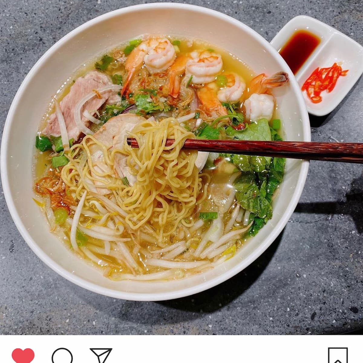 A bowl of noodle soup topped with shrimp, slices of meat, greens, and bean sprouts, with chopsticks resting on the bowl. This flavorful soup could easily rank among the best soup in every state. Sliced red chili and soy sauce sit beside it on a gray surface.