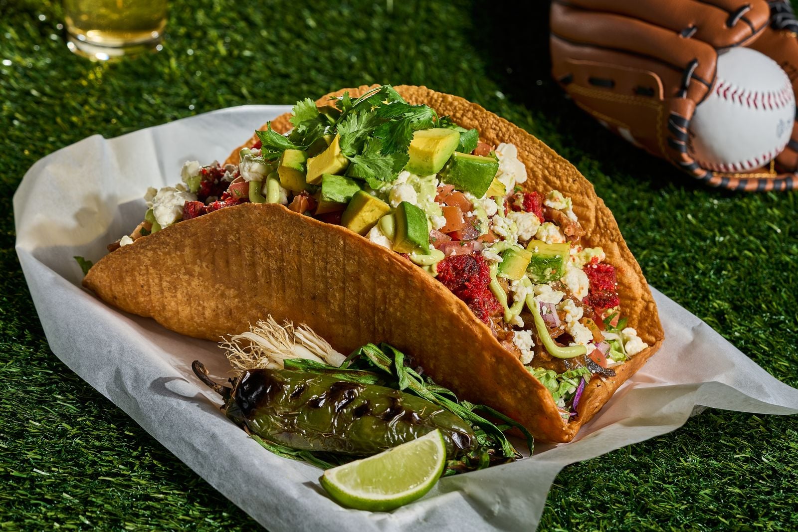 A crispy taco shell filled with avocado, cheese, lettuce, tomatoes, and meat sits on parchment paper with a grilled pepper, herbs, and lime wedges; a baseball and glove are in the background on green turf.